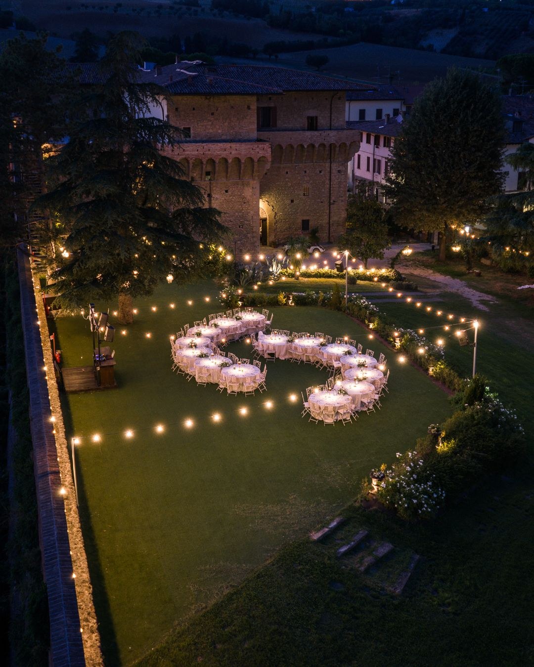 Tables arranged in a curve on the lawn at night, outlined with lights, Castello del Capitano delle Artiglierie