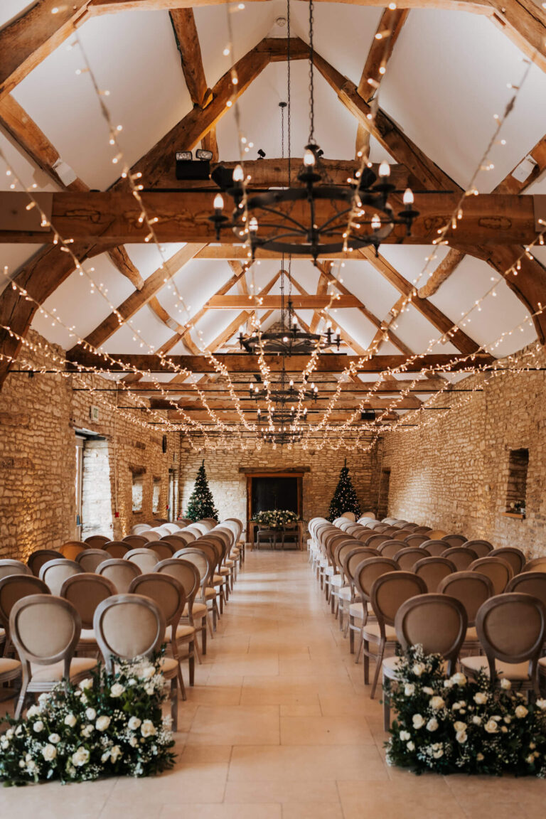 Rows of Louis chairs either side of the aisle in the barn, under iron chandeliers and fairy lights, Caswell House