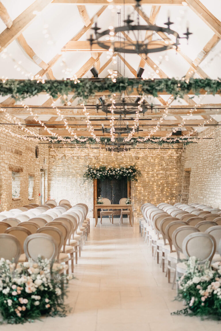 The barn ceremony aisle with white flowers, a garland of greenery and fairy lights across the beams, Caswell House