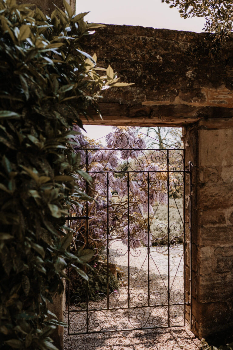 A wrought-iron gate in a stone wall, with wisteria in the garden beyond, Caswell House