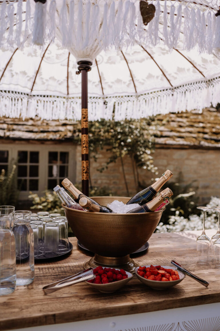 A champagne bucket and glasses on a courtyard drinks table under a tasselled parasol, Caswell House