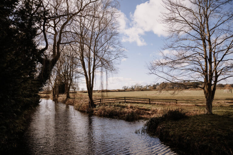 The moat edged by bare trees, with fields beyond, on a clear autumn day, Caswell House