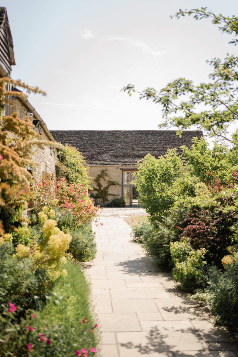 A paved path between flowering borders leading to one of the barns, Caswell House