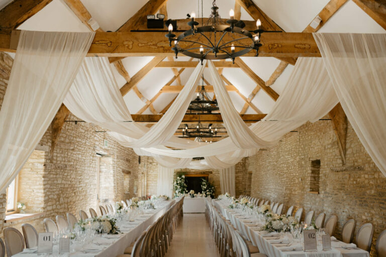 Long tables set for a wedding breakfast in the barn, with white fabric draped from the roof beams, Caswell House