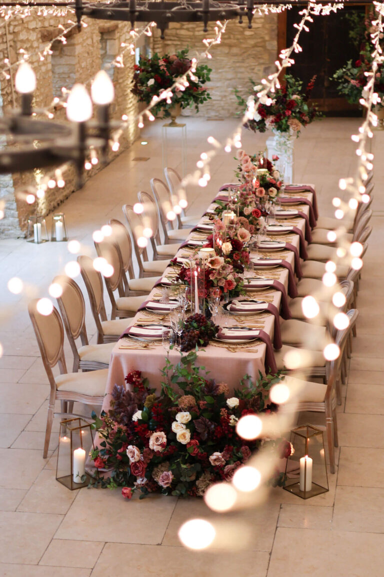 A long table dressed for a winter wedding, with deep red flowers, candle lanterns and strings of lights, Caswell House