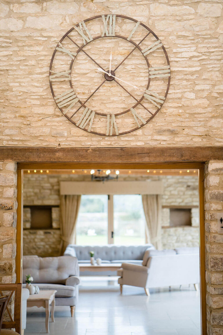 A large clock on the stone wall above a doorway between the barns, with a lounge area beyond, Caswell House