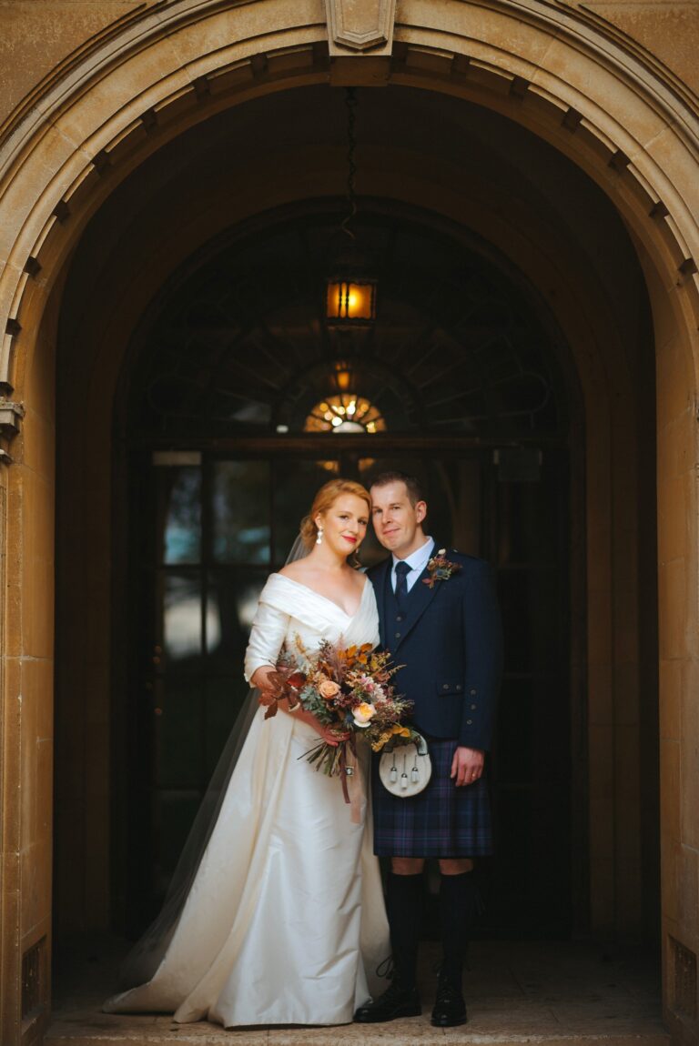 A bride in an ivory gown with full sleeves and a groom in a kilt in a stone arched doorway, Catherine Blades Couture