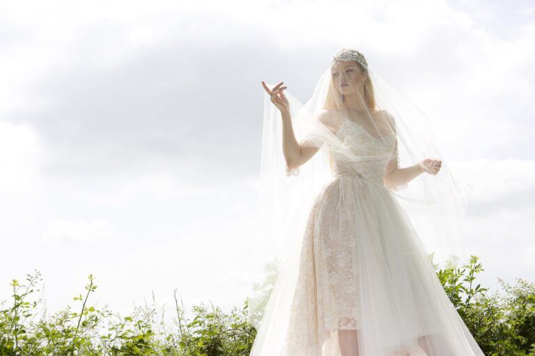 A bride in a lace-bodice gown with a long sheer veil against a pale sky, Catherine Blades Couture