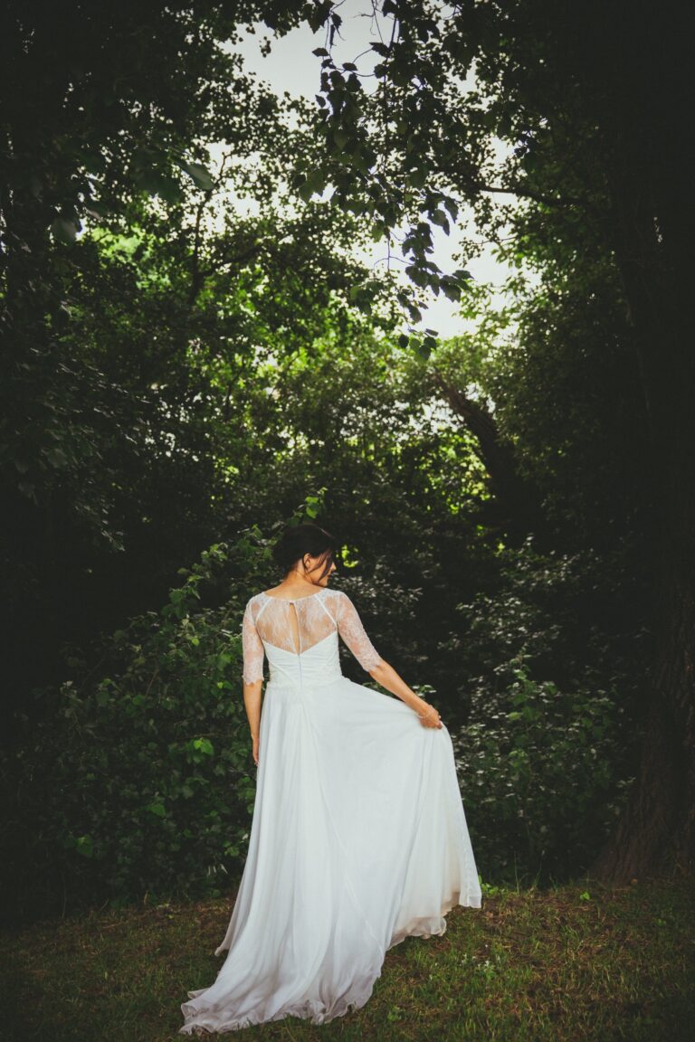 A bride in a lace-sleeved ivory gown lifting her skirt among trees, Catherine Blades Couture