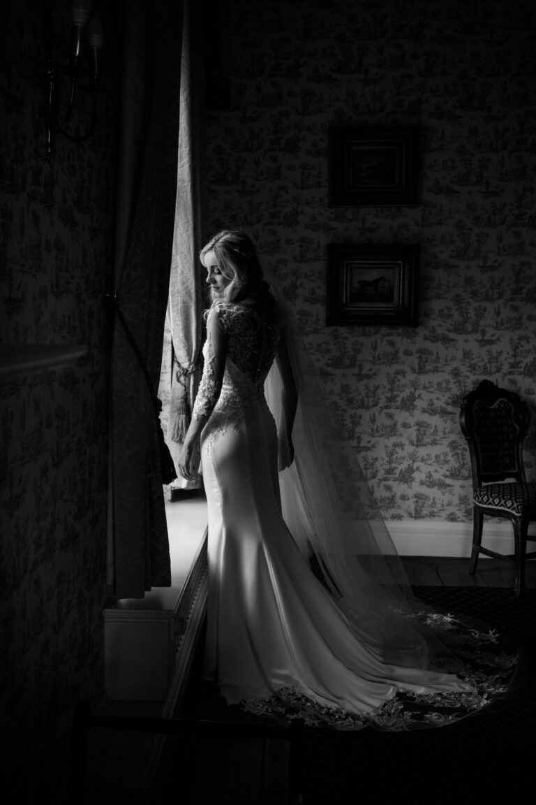 A black and white image of a bride by a window in a fitted gown with a long train, Catherine Blades Couture