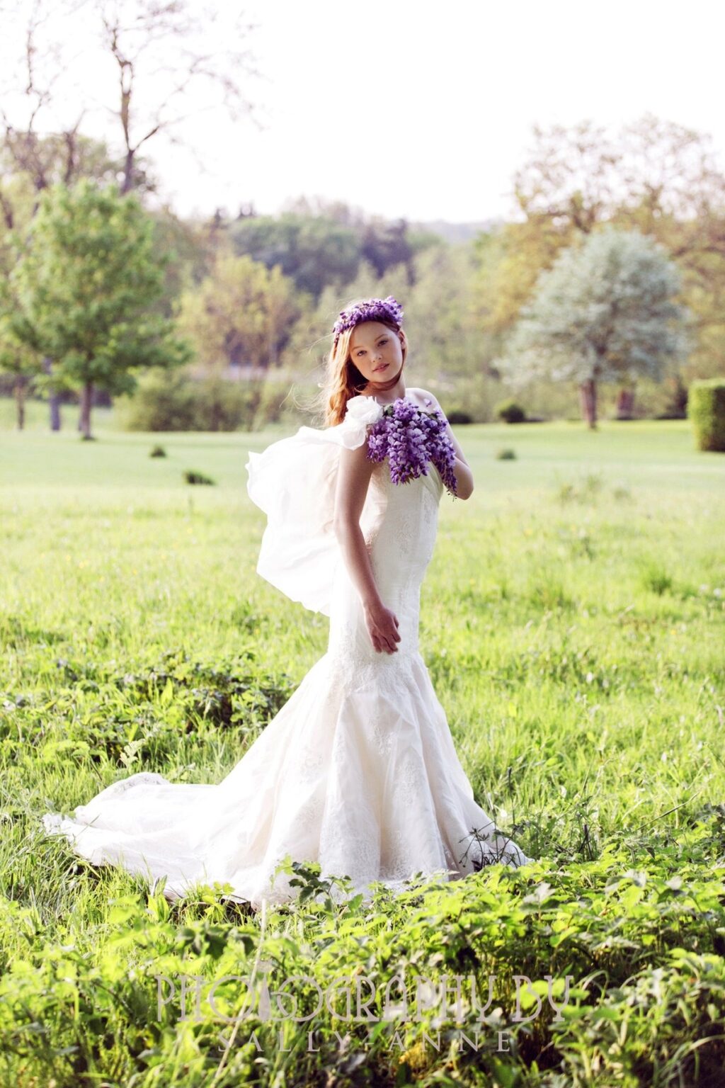 A bride in a fitted fishtail gown holding a purple bouquet on a lawn, Catherine Blades Couture