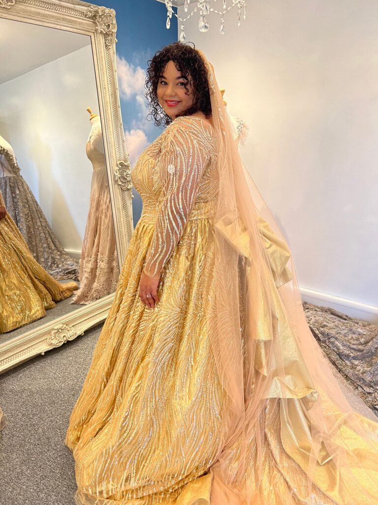 A client in a gold embellished gown with a long veil in the studio mirror room, Catherine Blades Couture