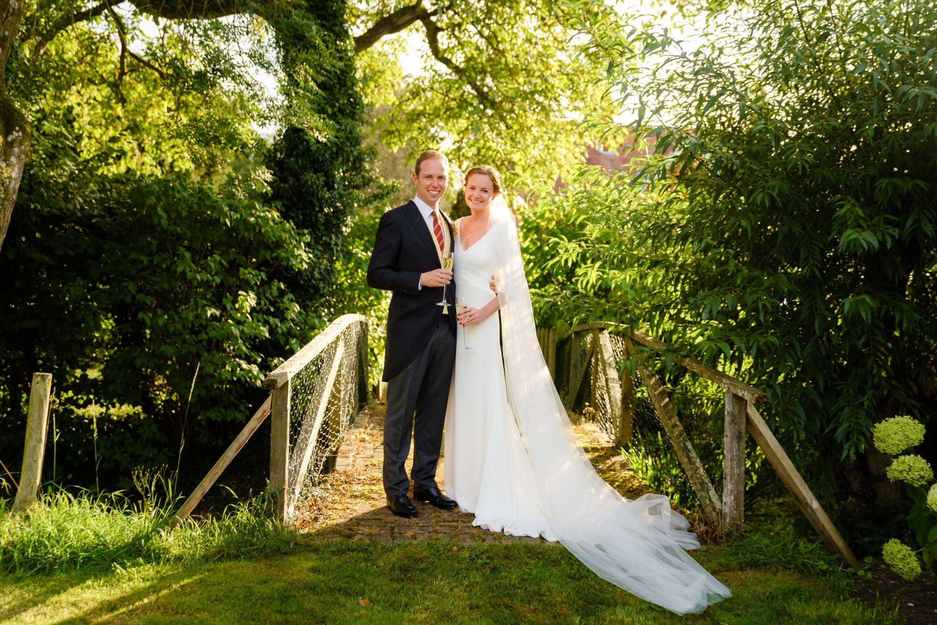 A bride in an ivory gown with a long veil beside her groom at a garden footbridge, Catherine Blades Couture