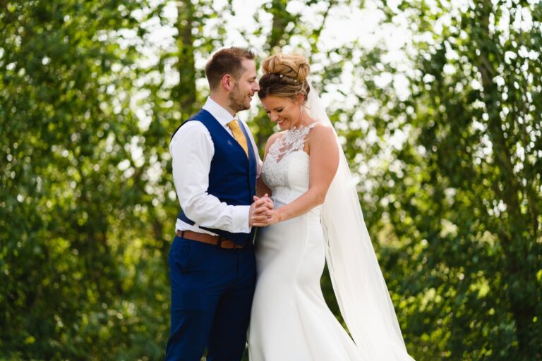 A couple holding hands among trees, the bride in a fitted gown with lace neckline and a long veil, Catherine Blades Couture