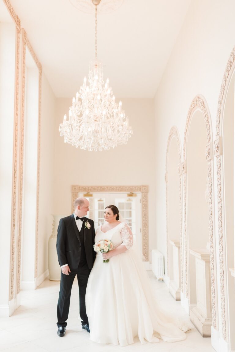 A bride in a ball gown and groom in black tie in a pale hall with a chandelier, Catherine Blades Couture