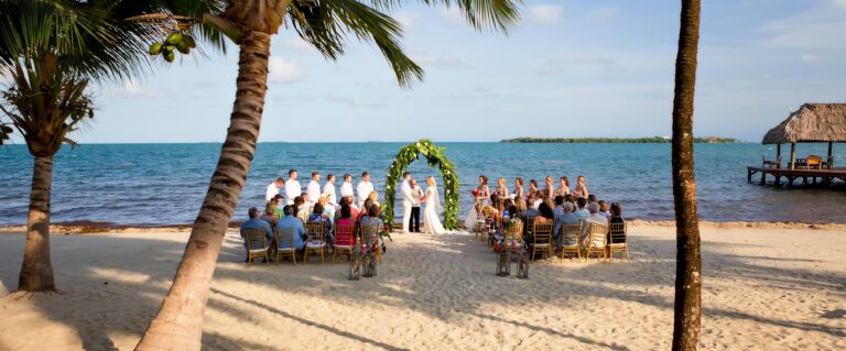 A beach wedding ceremony set on the sand, a floral arch and seated guests with the Caribbean behind, Chabil Mar Resort