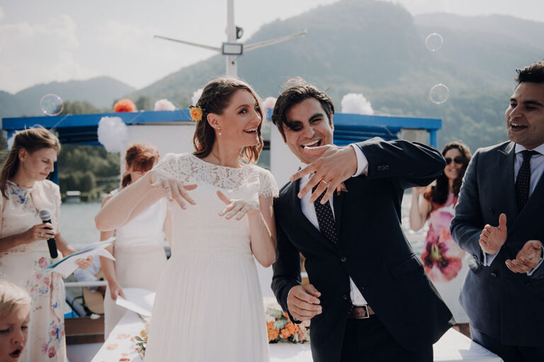 Bride and groom laughing on a lake boat with guests and mountains behind, Cherry On Top