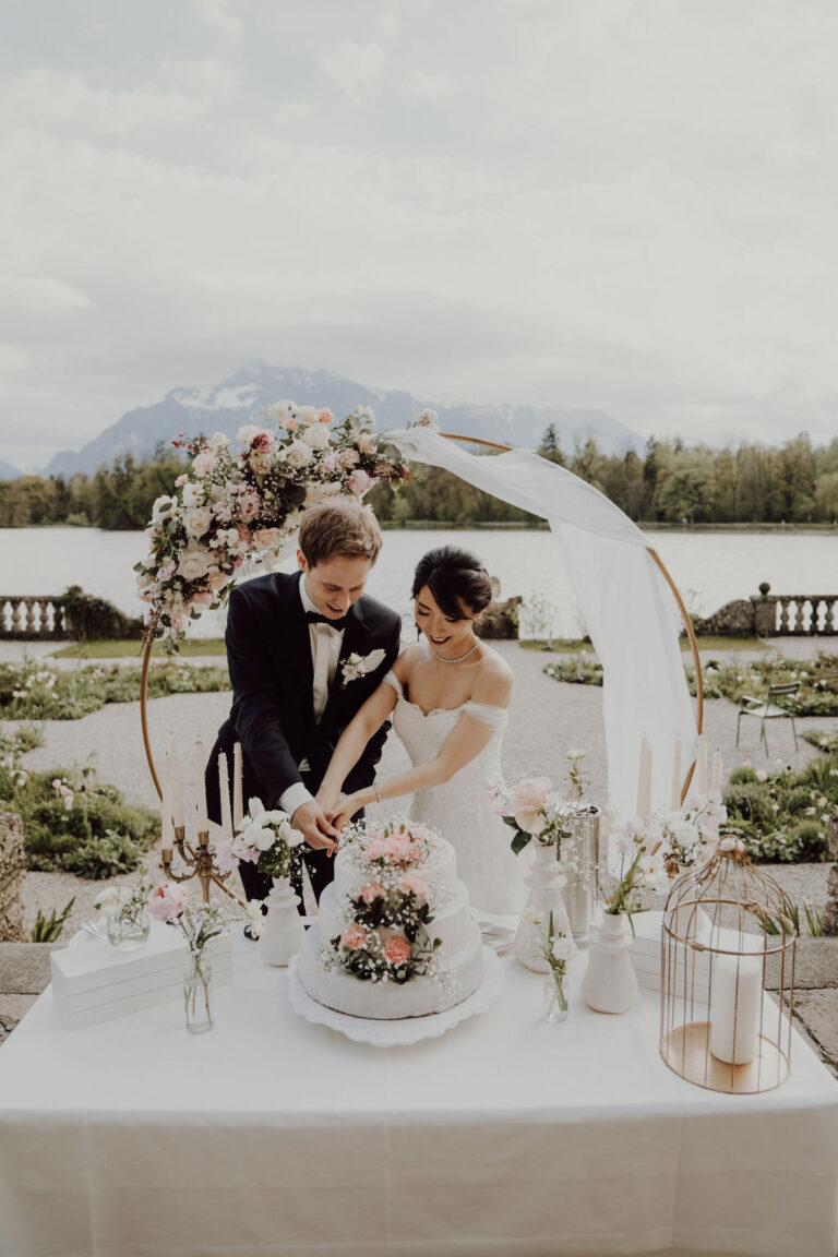 Couple cutting the cake at a table with a floral ring arch overlooking a lake, Cherry On Top