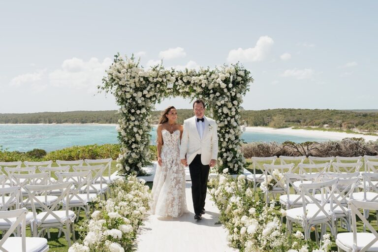 Couple walking through a white floral arch on a headland ceremony aisle above the sea, Chic Bahamas Weddings