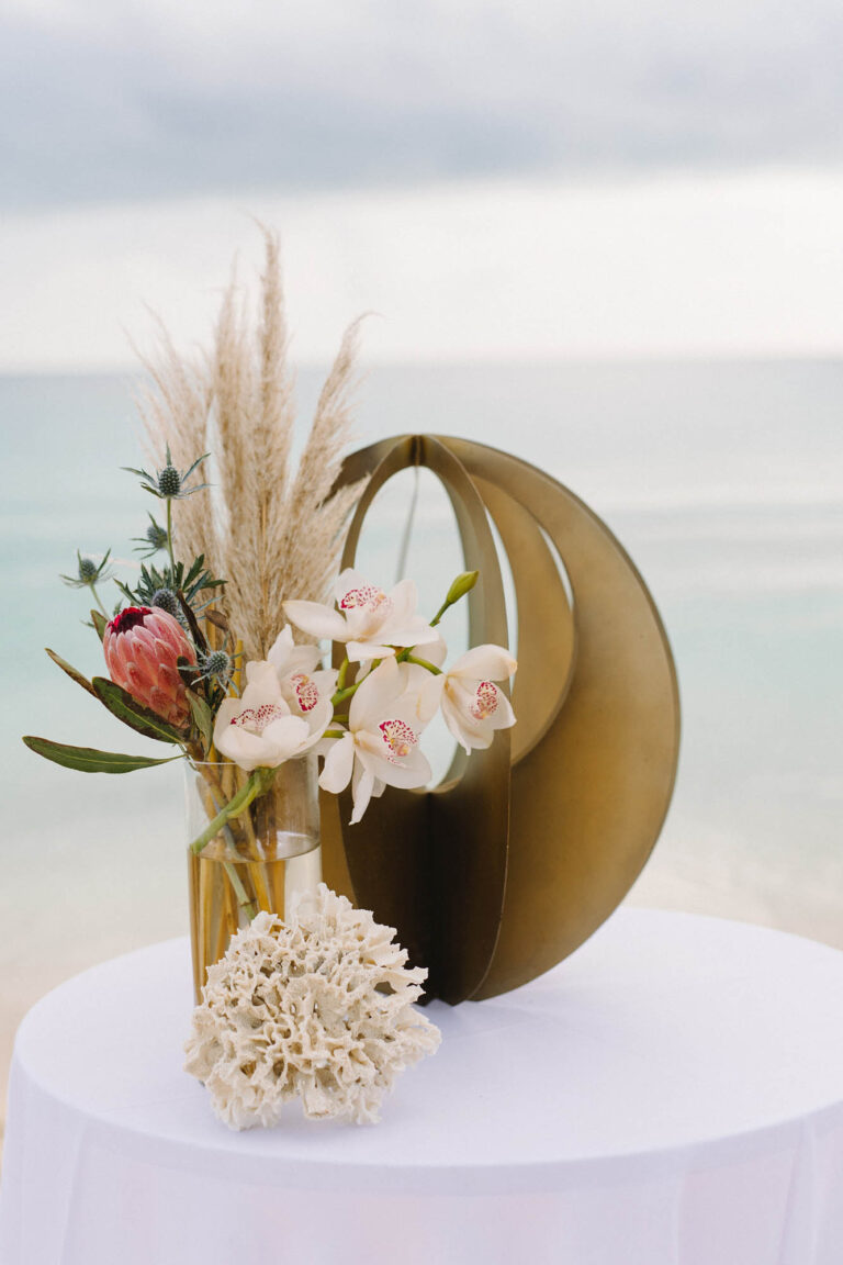 Modern sculpture with pampas grass, protea and coral on a white-clothed table by the sea, Chic Bahamas Weddings