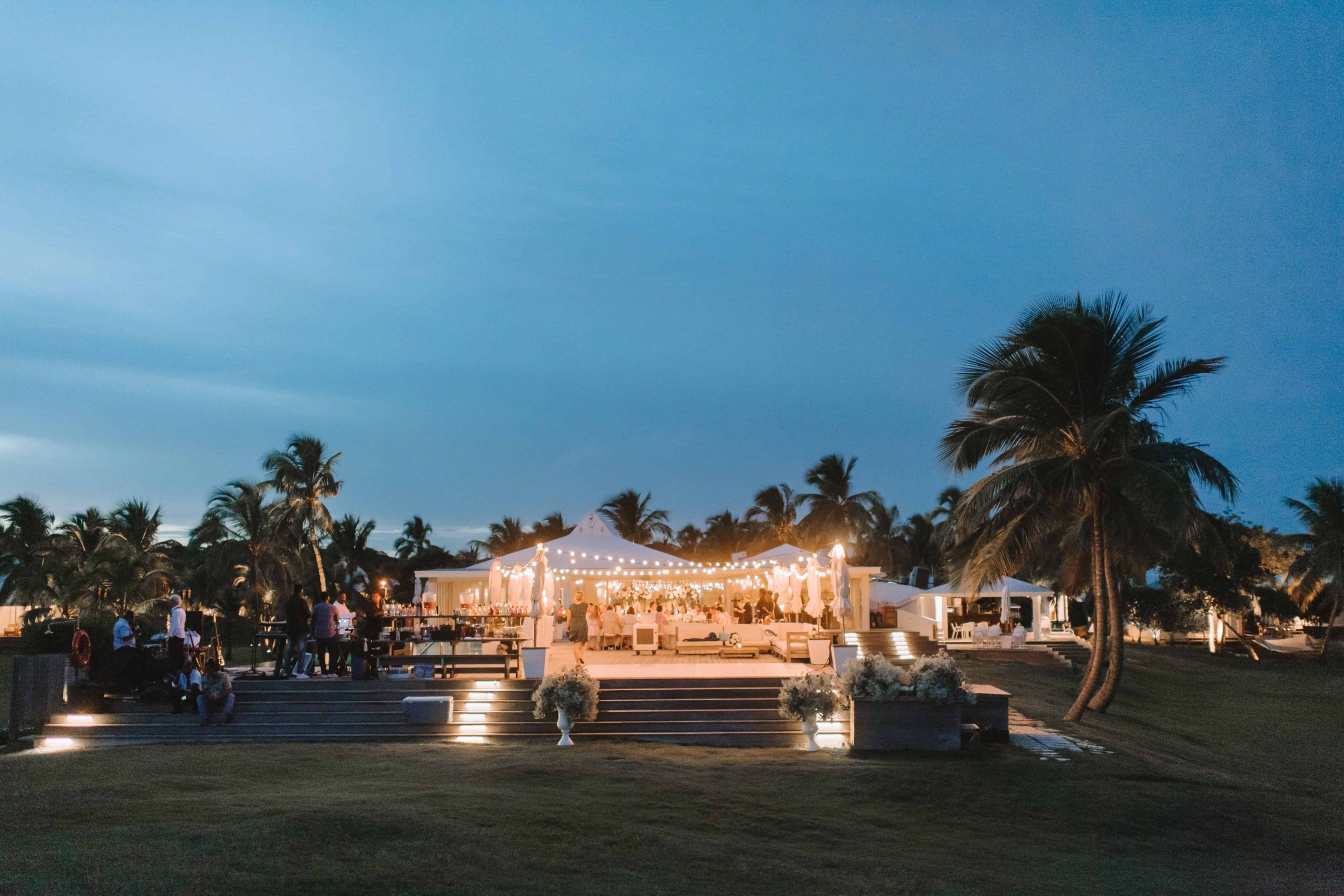 Reception tent lit up at dusk on a lawn with palms against a blue sky, Chic Bahamas Weddings