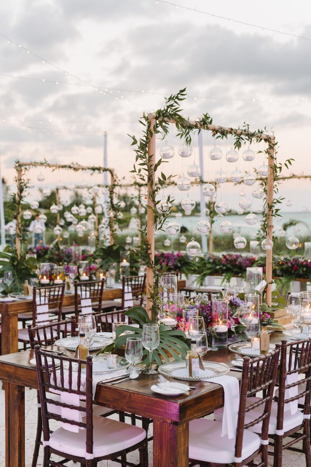 Dining tables on a beach at dusk, set between frames of hanging glass globes