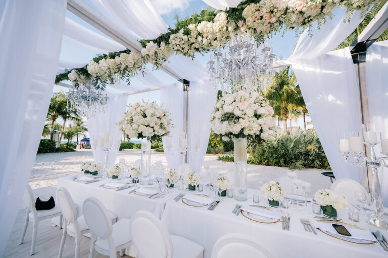 Reception table under a clear tent hung with white drapes and floral garlands, Chic Bahamas Weddings
