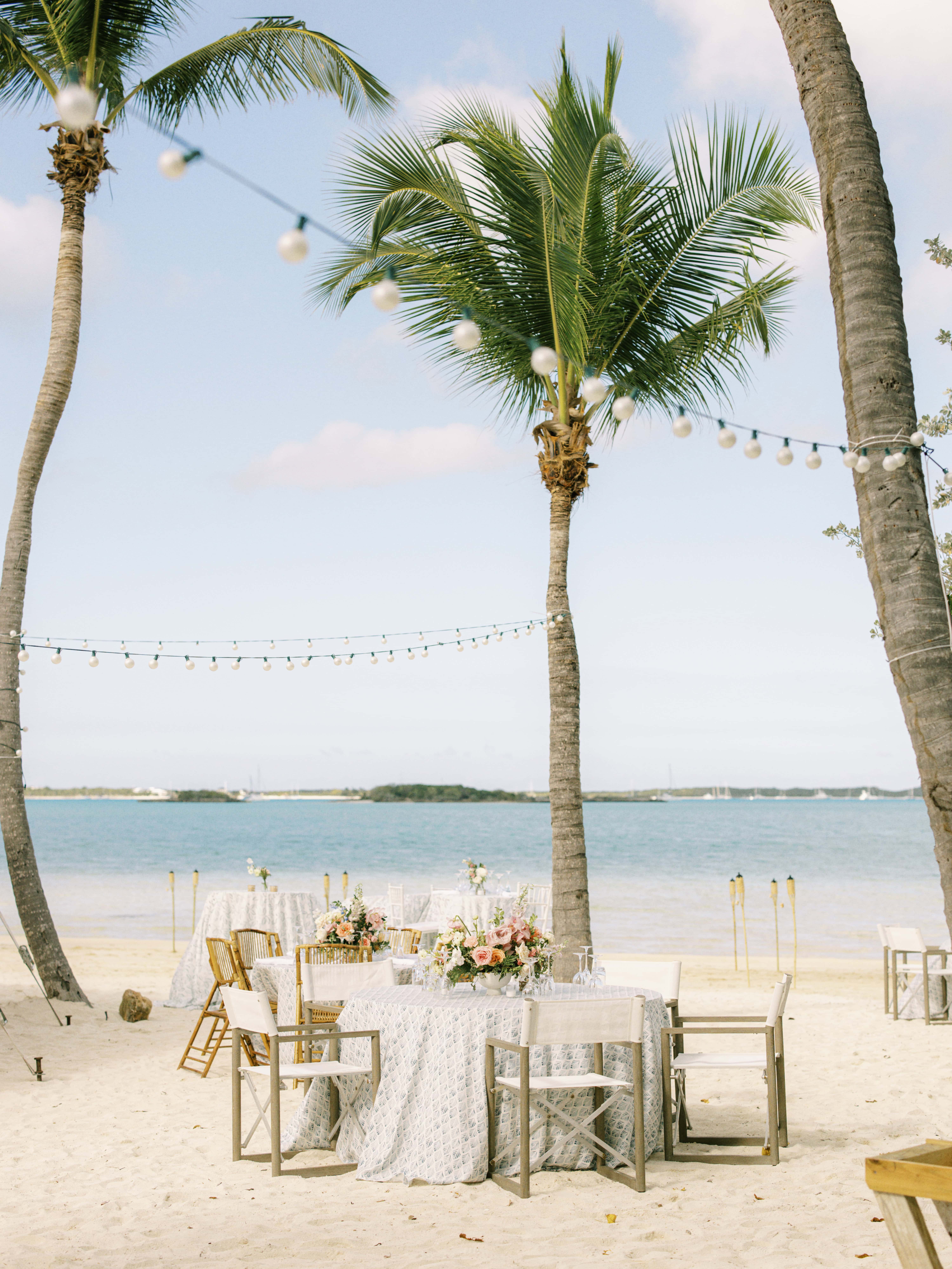 Beach dinner table set between palm trees under festoon lights, overlooking the water, Chic Bahamas Weddings