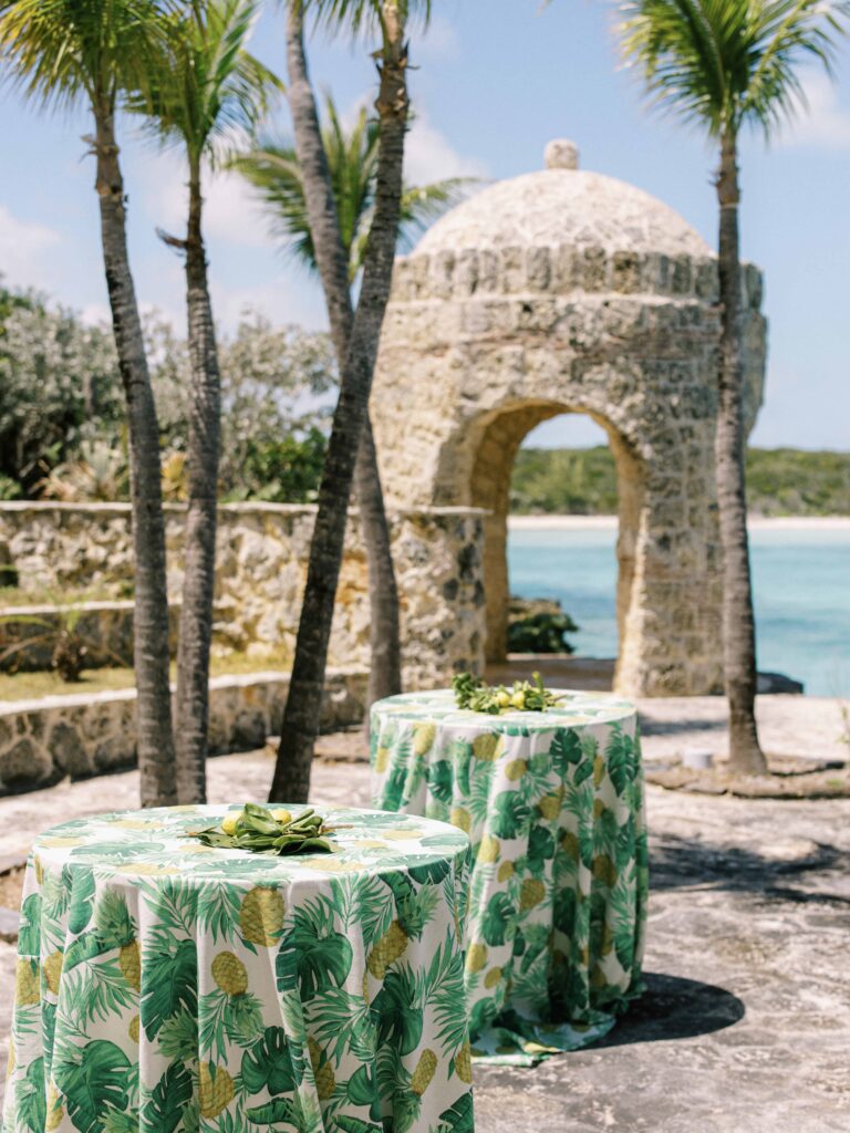 Cocktail tables in palm-print cloths beside a stone lookout with turquoise sea behind, Chic Bahamas Weddings