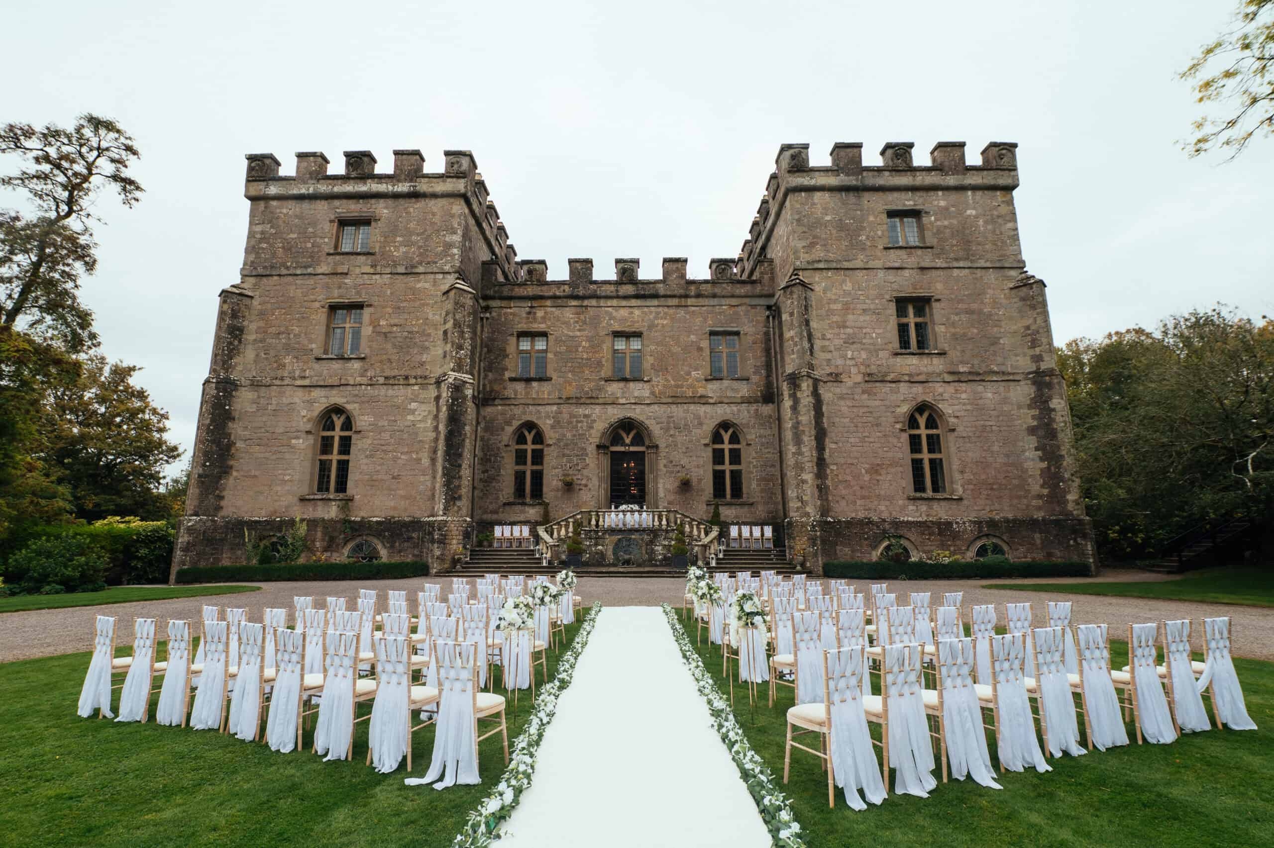 The castle's castellated front facade above a lawn aisle of white draped chairs set for an outdoor ceremony, Clearwell Castle