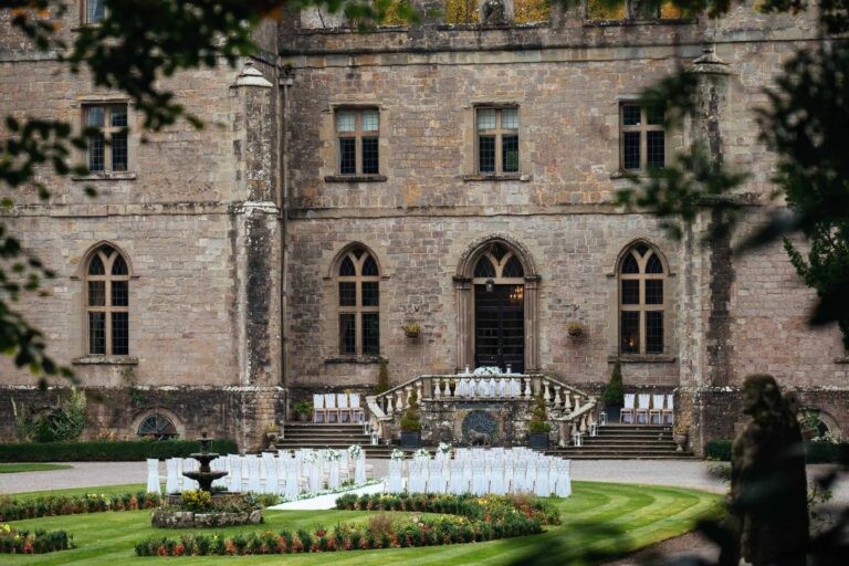 The rear elevation and balustraded terrace steps above a formal garden set with chairs for a ceremony, a fountain on the lawn, Clearwell Castle