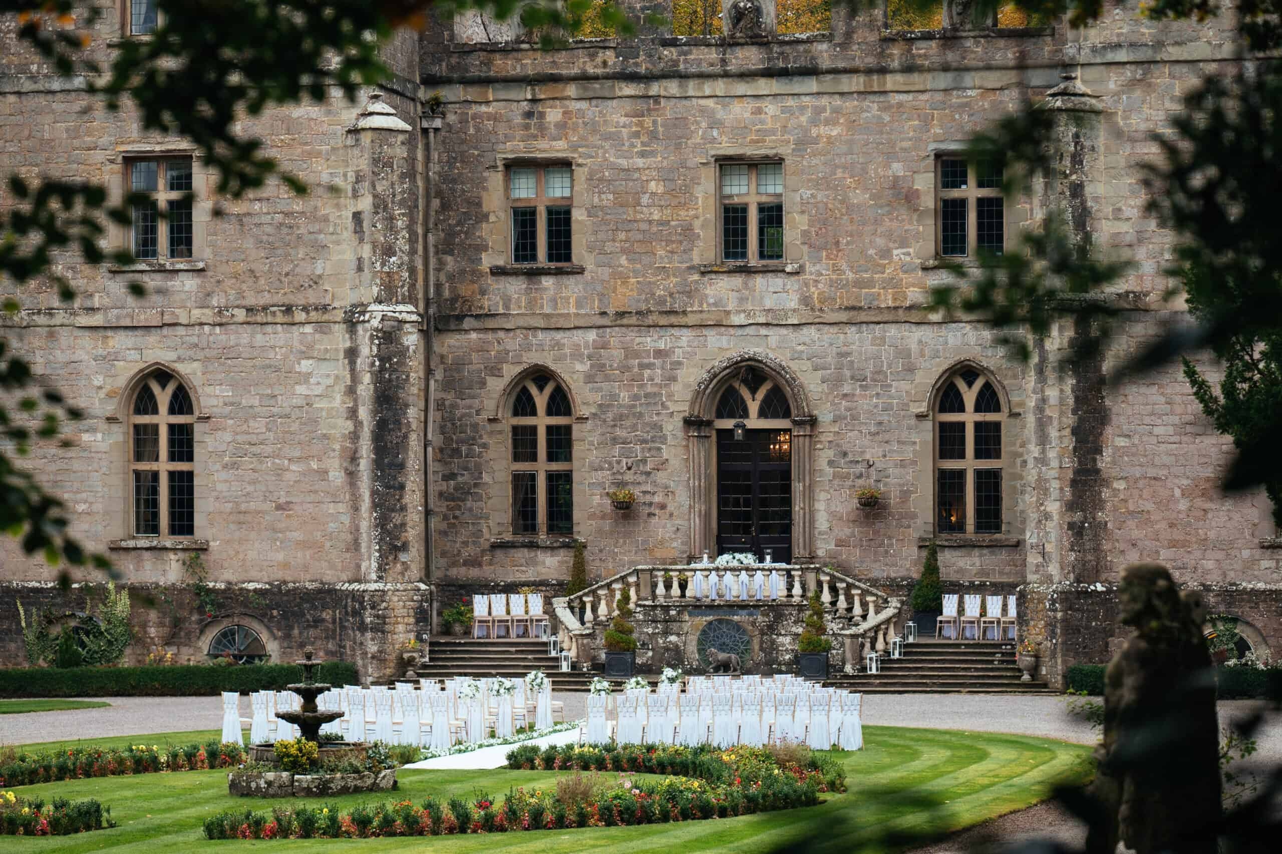 The rear elevation and balustraded terrace steps above a formal garden set with chairs for a ceremony, a fountain on the lawn, Clearwell Castle