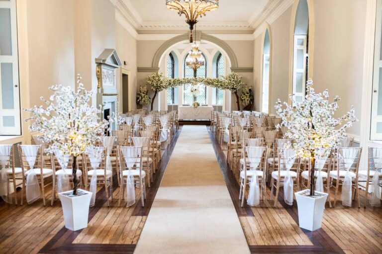 A ceremony in the Ballroom, blossom trees flanking the aisle beneath the chandelier and arched leaded window, Clearwell Castle