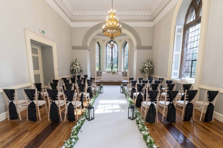 A Ballroom ceremony dressed with dark chair sashes, the aisle lit by lanterns to the arched windows, Clearwell Castle