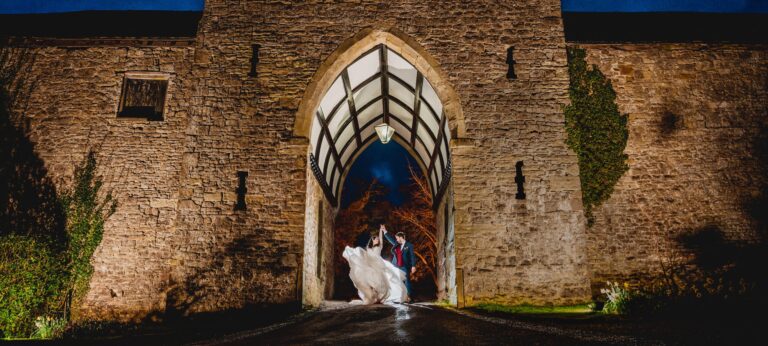 A couple framed in the lit gatehouse arch at dusk, the medieval gateway glowing, Clearwell Castle