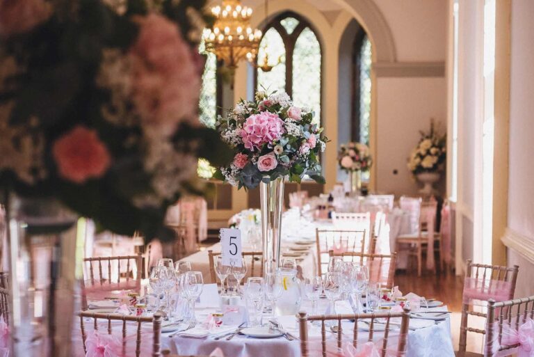 The Ballroom laid for the wedding breakfast, a tall pink centrepiece above chiavari chairs and arched windows, Clearwell Castle