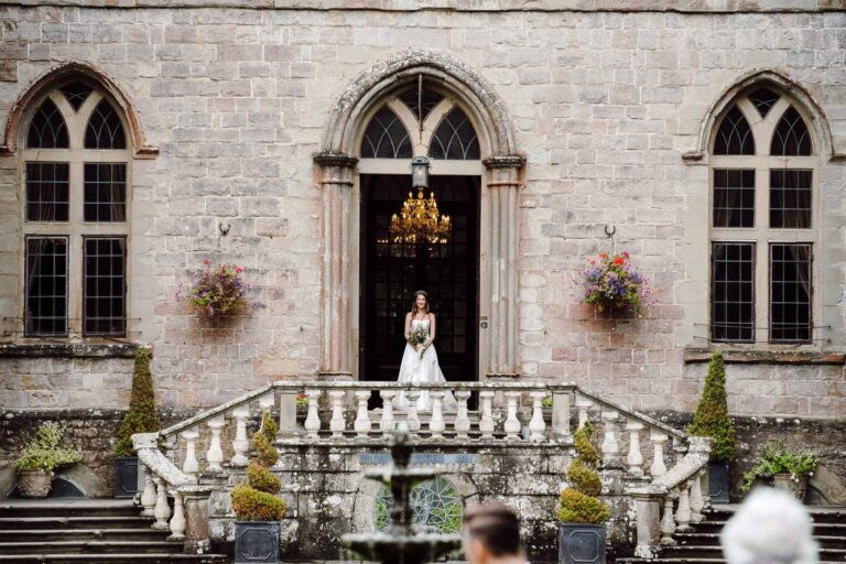 The bride on the balustraded steps at the castle's arched entrance, a chandelier glowing within, Clearwell Castle