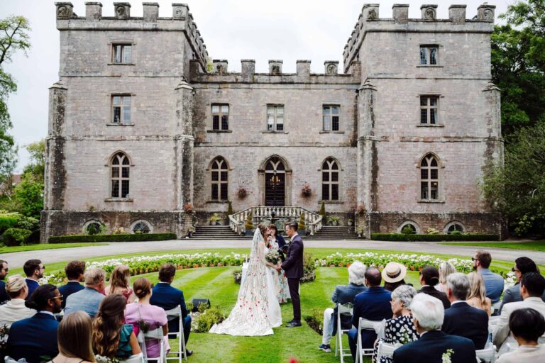 An outdoor ceremony on the lawn, the couple exchanging vows before the castle with guests seated around, Clearwell Castle