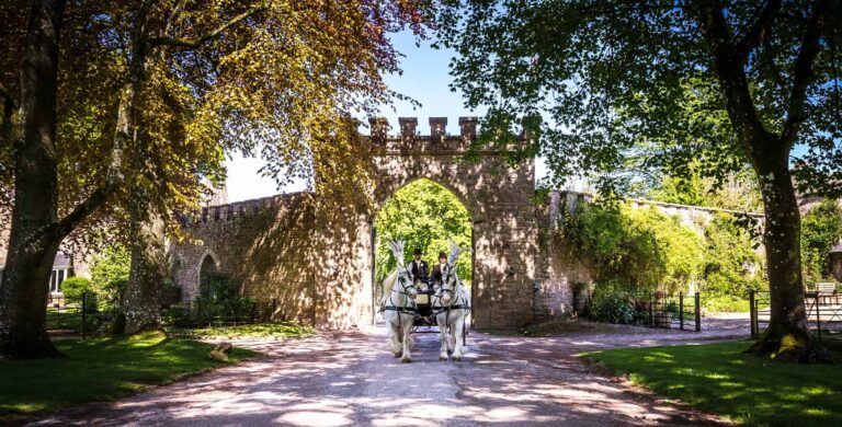 A white horse-drawn carriage arriving through the castle's gatehouse arch on a summer day, Clearwell Castle