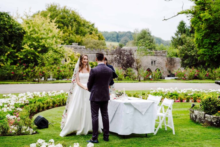 A ceremony by the garden Bandstand, the couple at the table amid flower beds and the castle wall beyond, Clearwell Castle
