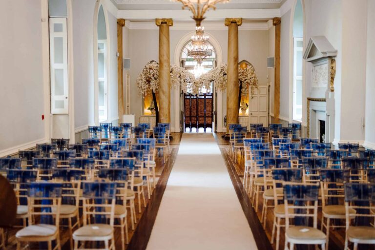 A ceremony in the columned Ballroom, blue chairs set to the arched entrance doors beneath the chandelier, Clearwell Castle