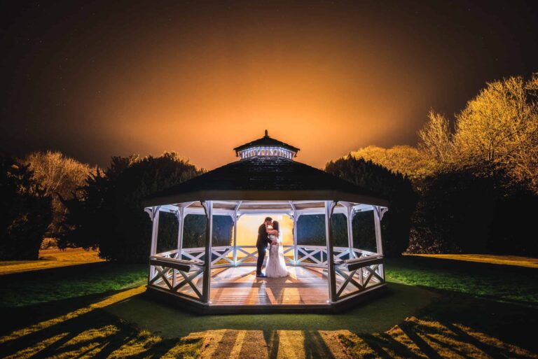 A couple embracing in the illuminated garden Bandstand at night, the sky burning amber behind, Clearwell Castle