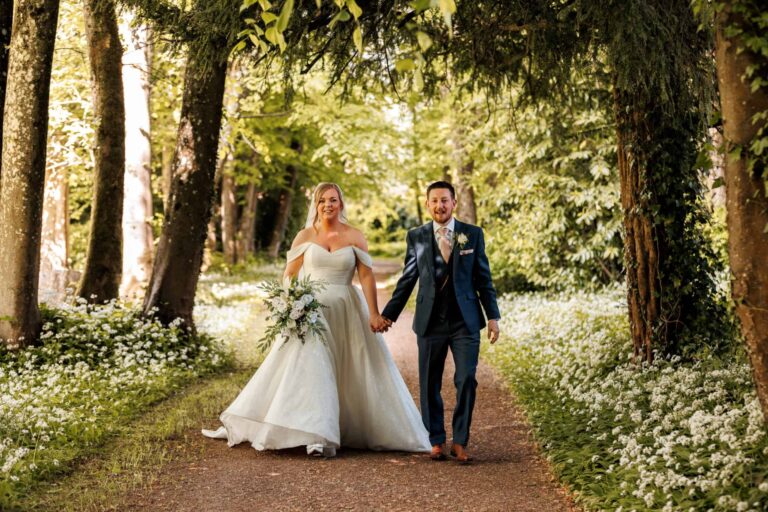 A couple walking a tree-lined path through the grounds, wild garlic in flower on either side, Clearwell Castle