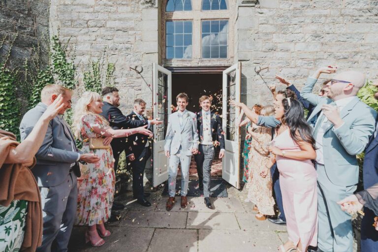 A confetti send-off, the couple stepping from the castle doorway through a shower thrown by guests, Clearwell Castle