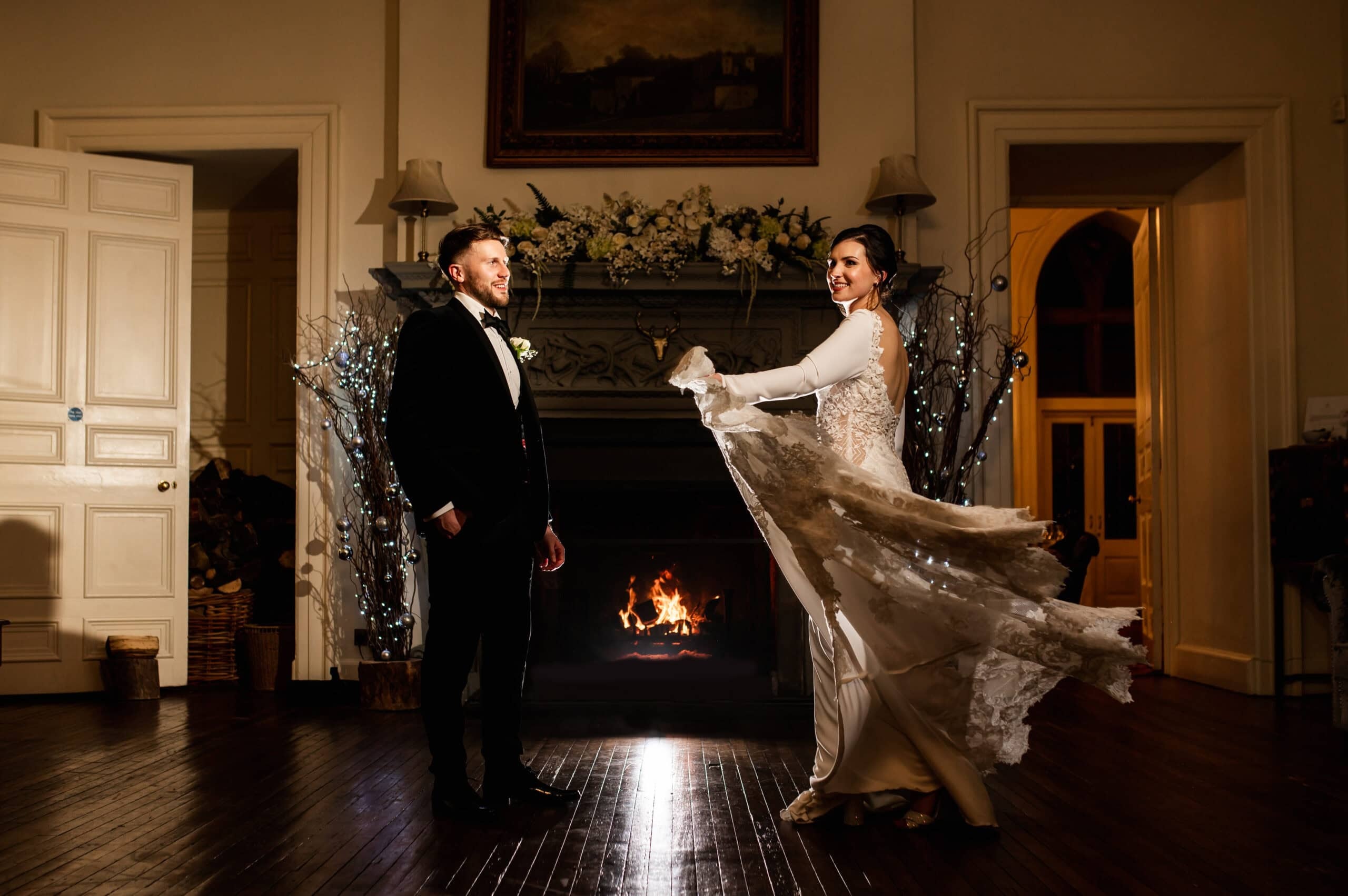 A couple by a blazing fireplace in a candlelit reception room, the bride's gown caught mid-turn, Clearwell Castle