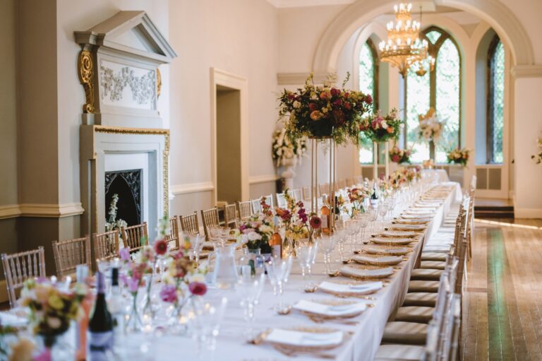 A long banquet table dressed for the wedding breakfast, tall florals and candlelight beneath the chandelier, Clearwell Castle