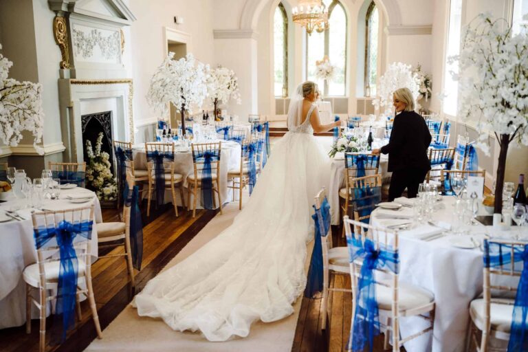 The bride crossing the Ballroom in a long train, blue chair sashes and blossom for the reception, Clearwell Castle