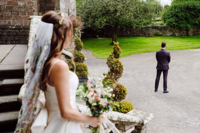 A first look in the gardens, the bride with her bouquet by clipped topiary as the groom waits, Clearwell Castle