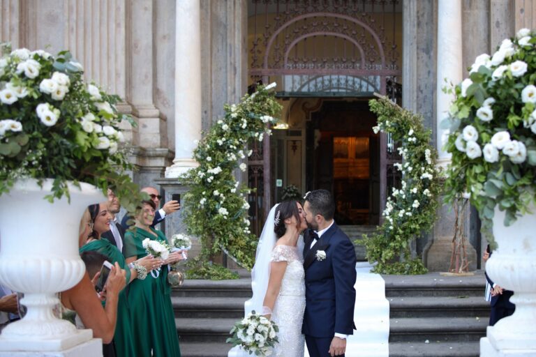 A kiss on the church steps between two arches of greenery and white flowers, with urns of white roses either side of the door, Clorinda Bellone Elite Weddings