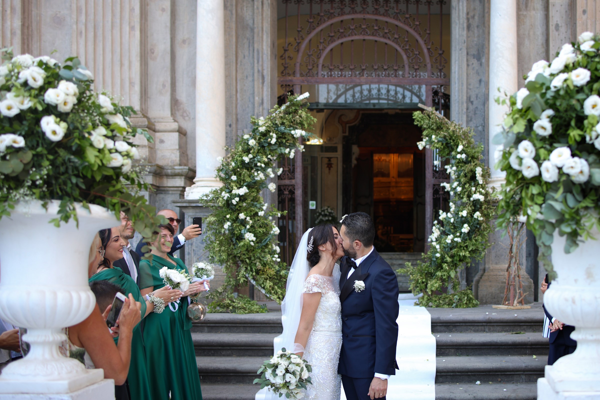 A kiss on the church steps between two arches of greenery and white flowers, with urns of white roses either side of the door, Clorinda Bellone Elite Weddings